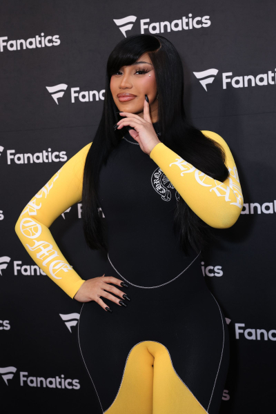Cardi B poses at an event in a black and yellow catsuit. Her hair is long and parted to the side. She wears black nail...
